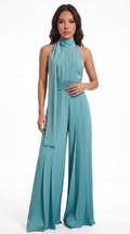 A woman modeling a sleeveless high-neck teal jumpsuit with a matching fabric tie at the waist, photographed against a plain white studio background, from Meleche Fashion USA or Meléche (short as (Mé))