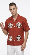 A man wearing a rust-orange short-sleeve button-up shirt with three large black and beige floral embroidered motifs on the front, paired with white pants, against a plain white background, from Meleche Fashion USA or Meléche (short as (Mé))