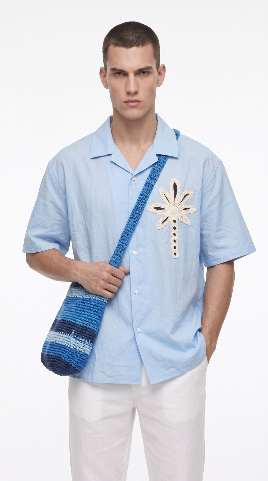A male model wearing a light blue short-sleeve button-up shirt with a palm tree patch on the chest, paired with white pants and a blue striped woven shoulder bag, from Meleche Fashion USA or Meléche (short as (Mé))