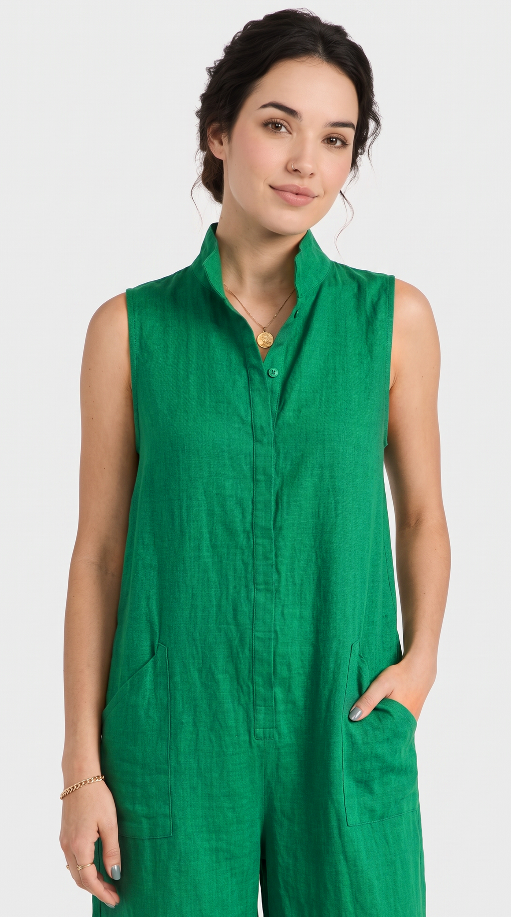 A woman wearing a sleeveless green linen jumpsuit with a stand-up collar and button front, photographed against a light gray studio background.