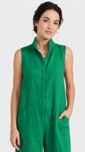A woman wearing a sleeveless green linen jumpsuit with a stand-up collar and button front, photographed against a light gray studio background.