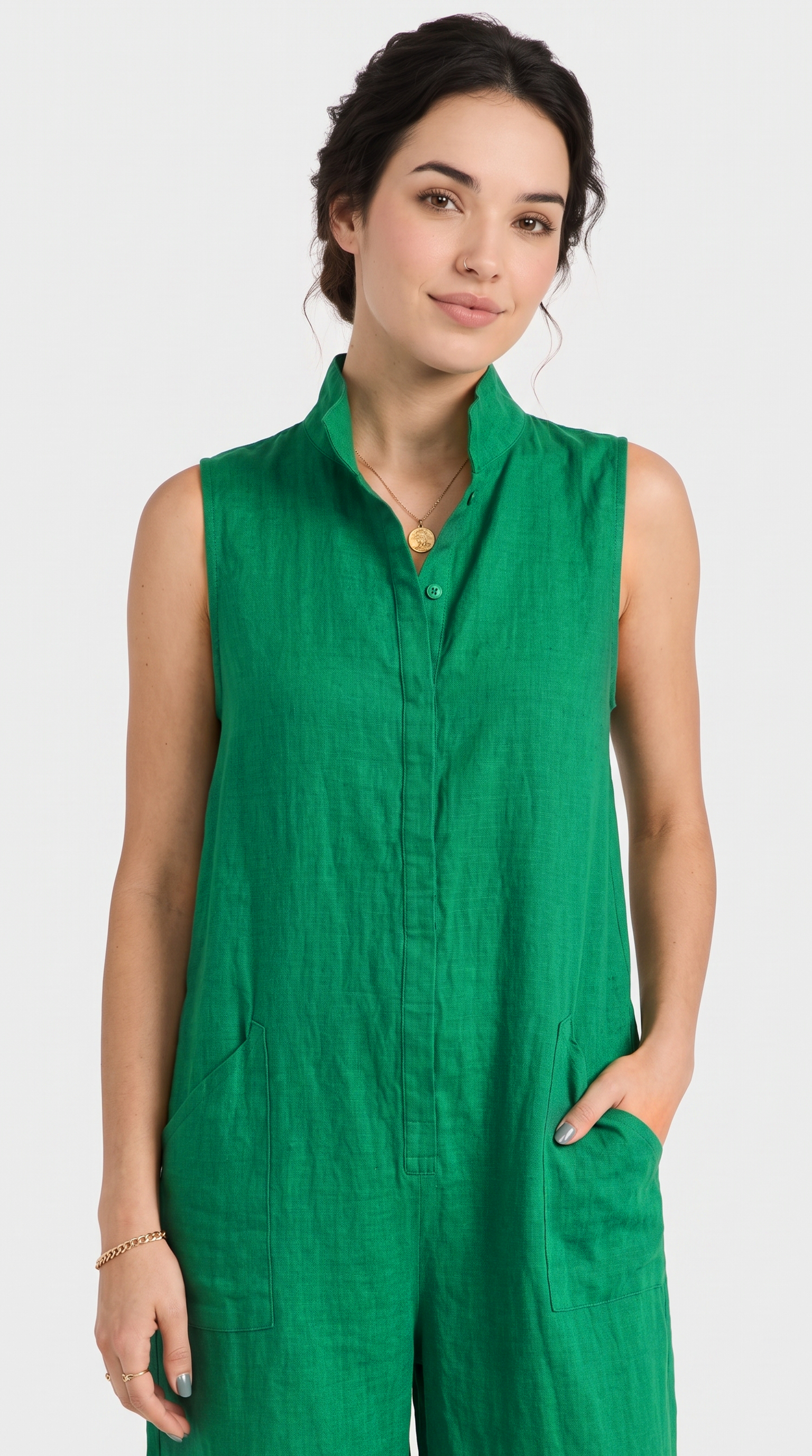 A woman wearing a sleeveless green linen jumpsuit with a stand-up collar and button front, photographed against a light gray studio background.