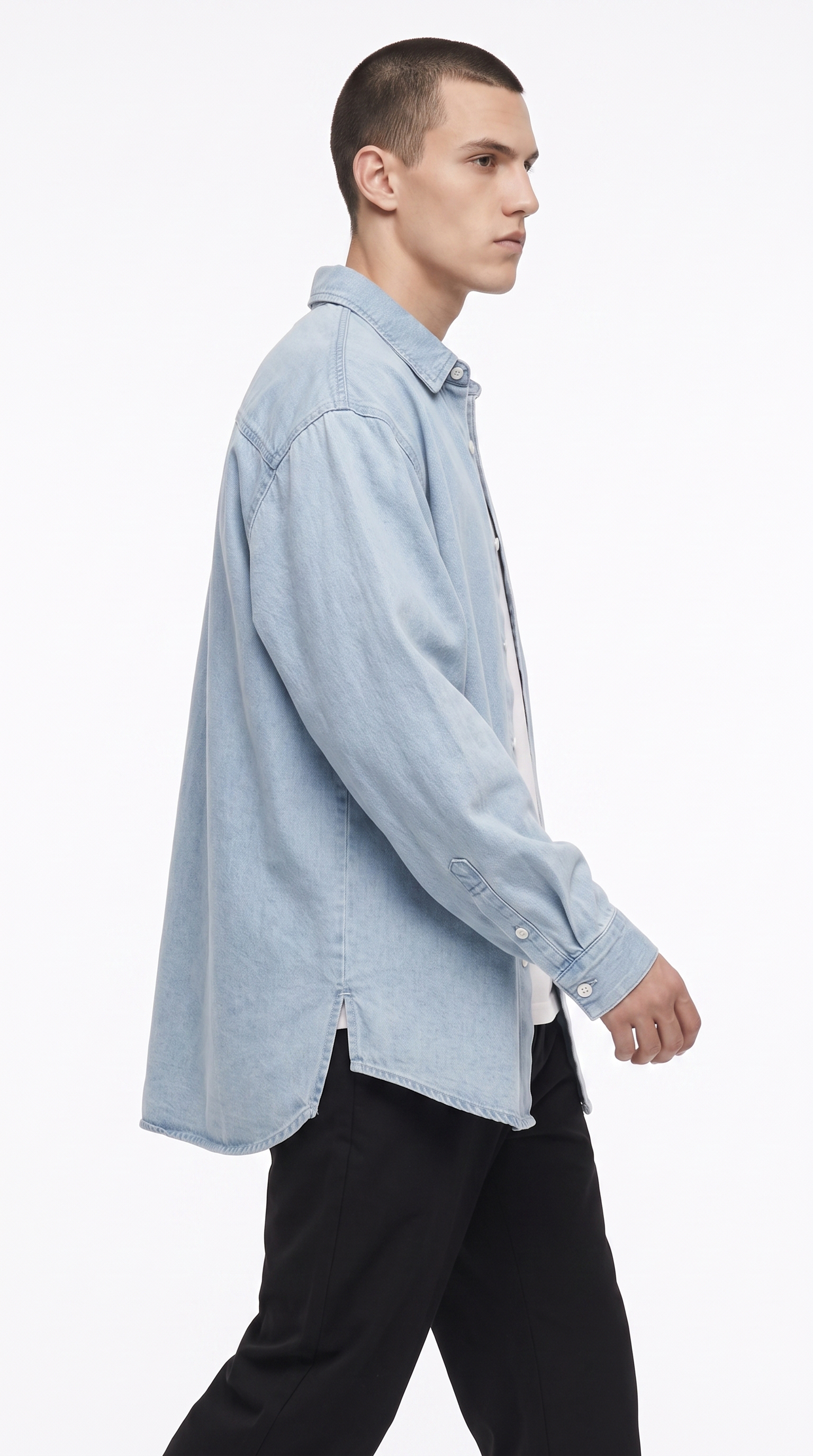 A man in profile wearing an oversized light blue denim shirt over a white undershirt and black pants, photographed against a clean white studio background, from Meleche Fashion USA or Meléche (short as (Mé))