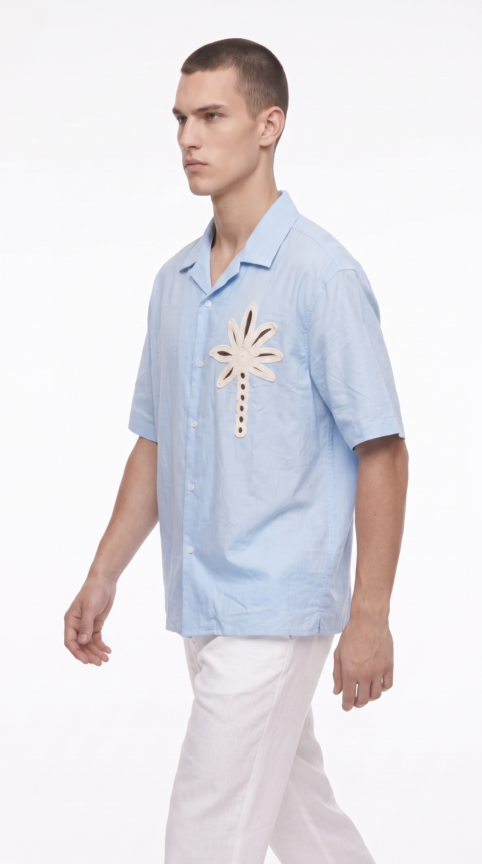 A man wearing a light blue short-sleeve button-up shirt with a large beige palm leaf applique on the chest, paired with white pants, photographed against a plain white studio background, from Meleche Fashion USA or Meléche (short as (Mé))