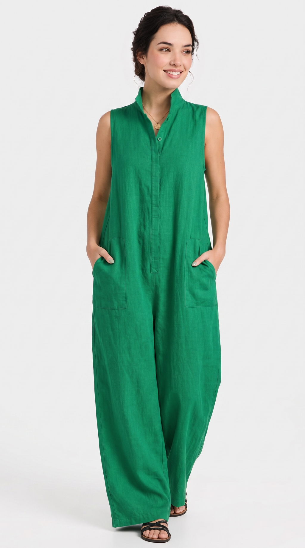 A woman modeling a sleeveless green jumpsuit with a high collar, front button placket, and side pockets, photographed against a plain light gray studio background.