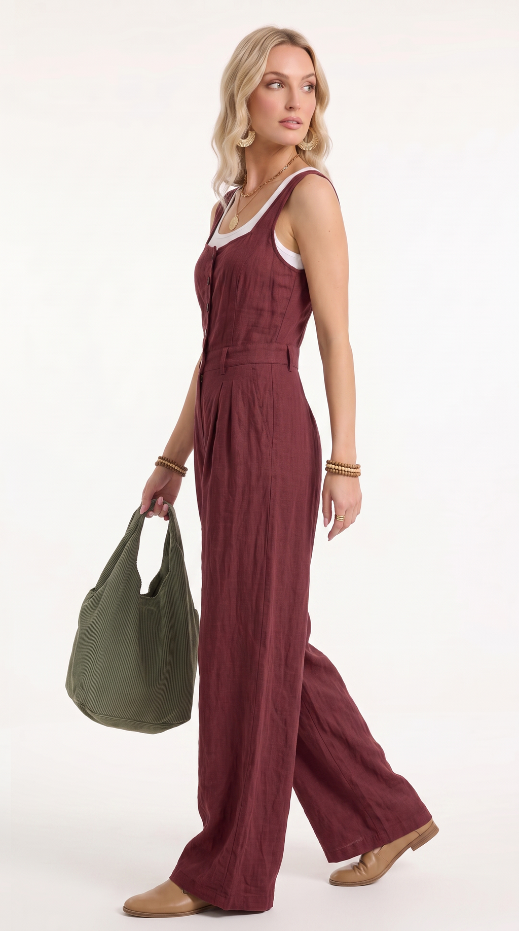 A woman modeling a sleeveless burgundy wide-leg jumpsuit with a white inner neckline, holding an olive green tote bag against a plain white studio background.