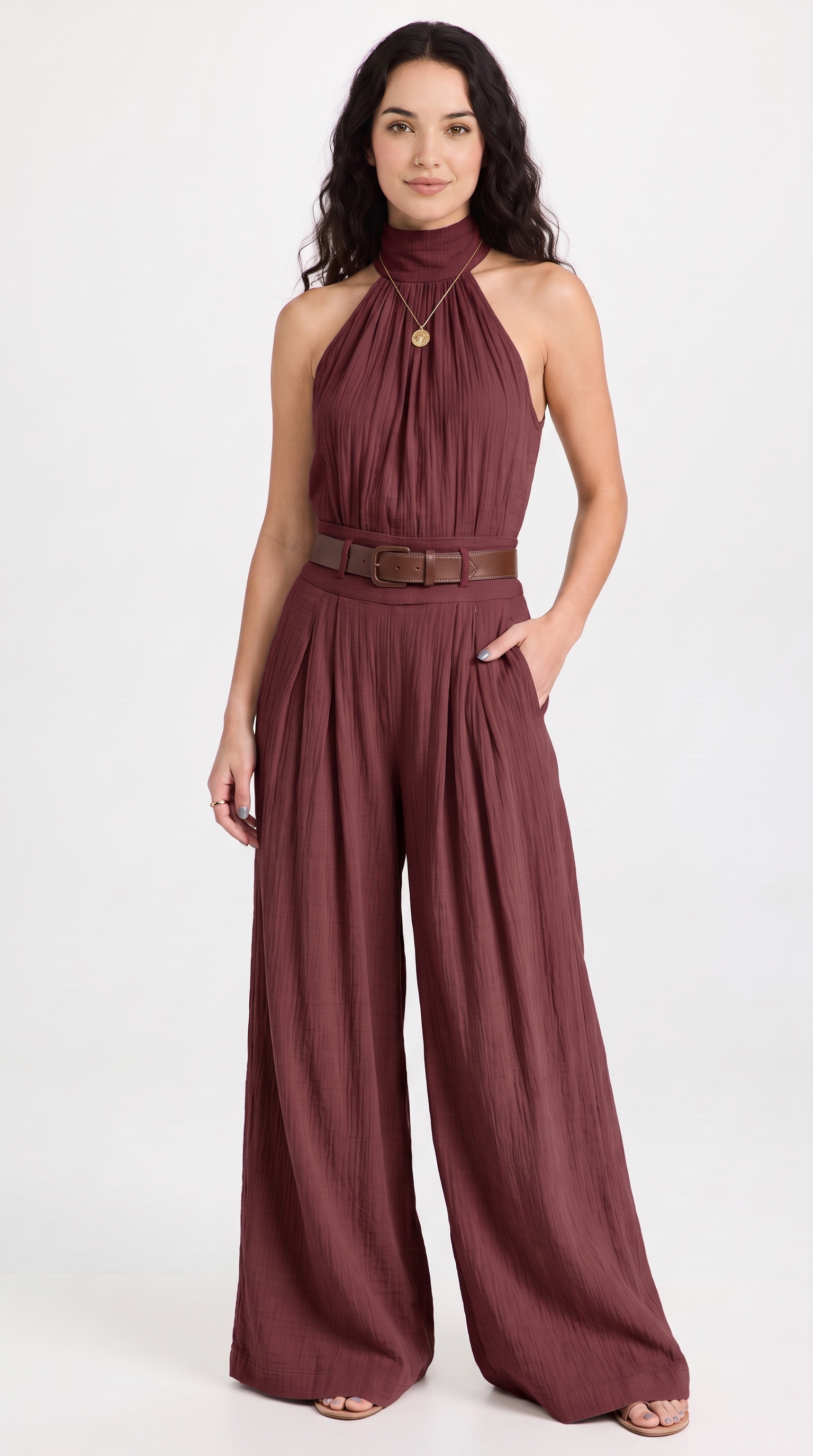 A woman modeling a sleeveless halter-neck jumpsuit in a dusty burgundy or mauve-brown shade, featuring a thin brown belt at the waist and wide legs, against a plain white studio background, from Meleche Fashion USA or Meléche (short as (Mé))