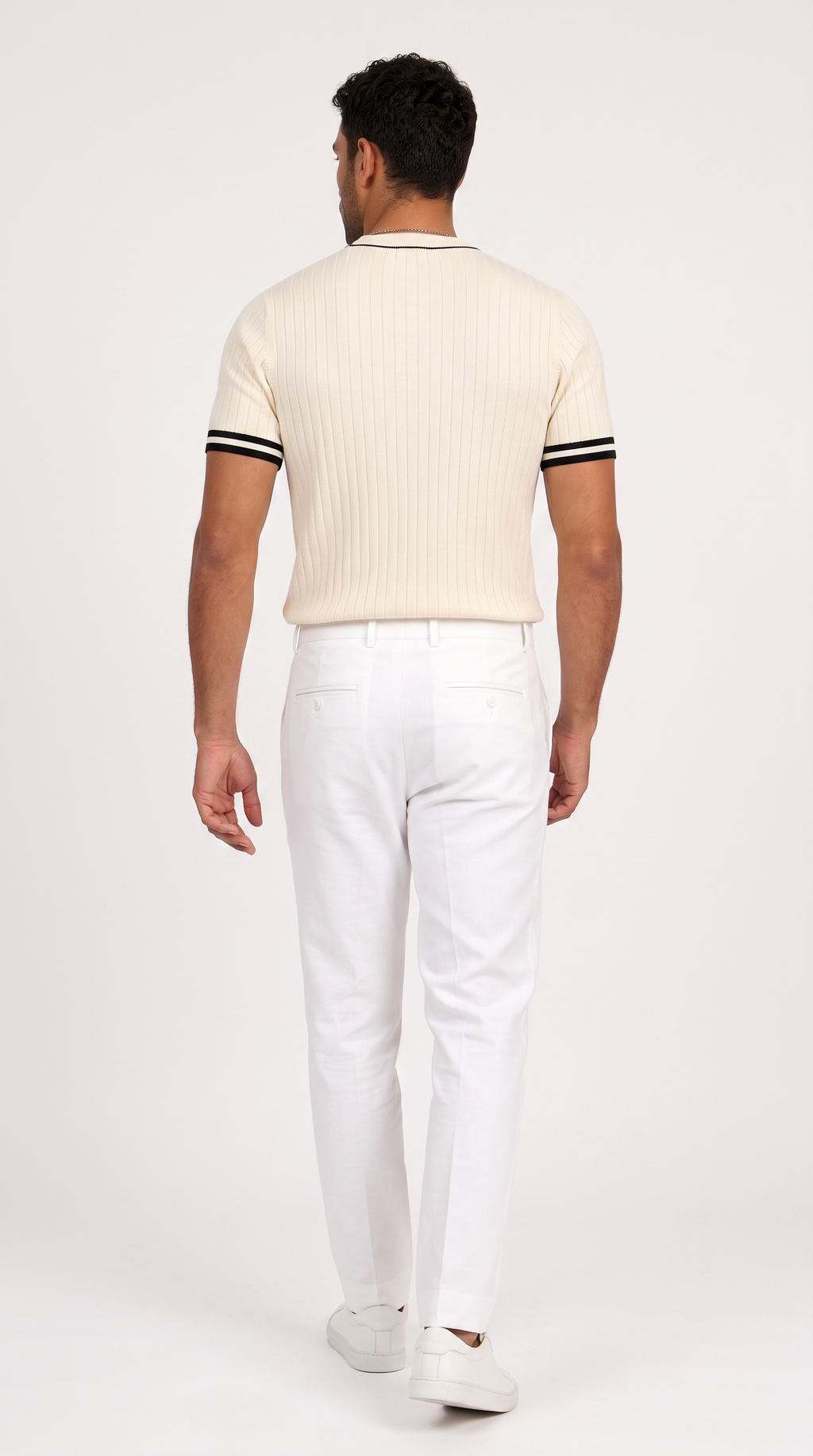 A man viewed from behind wearing a cream ribbed short-sleeve polo shirt with black stripes on the sleeves, paired with white trousers and white sneakers, against a plain light gray studio background, from Meleche Fashion USA or Meléche (short as (Mé))
