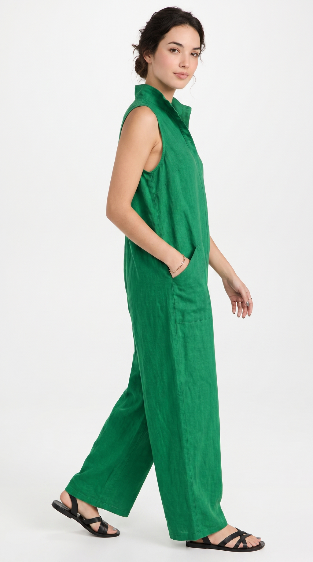 A woman modeling a sleeveless green wide-leg jumpsuit with a high collar and side pockets, paired with black strappy sandals, against a plain white studio background.