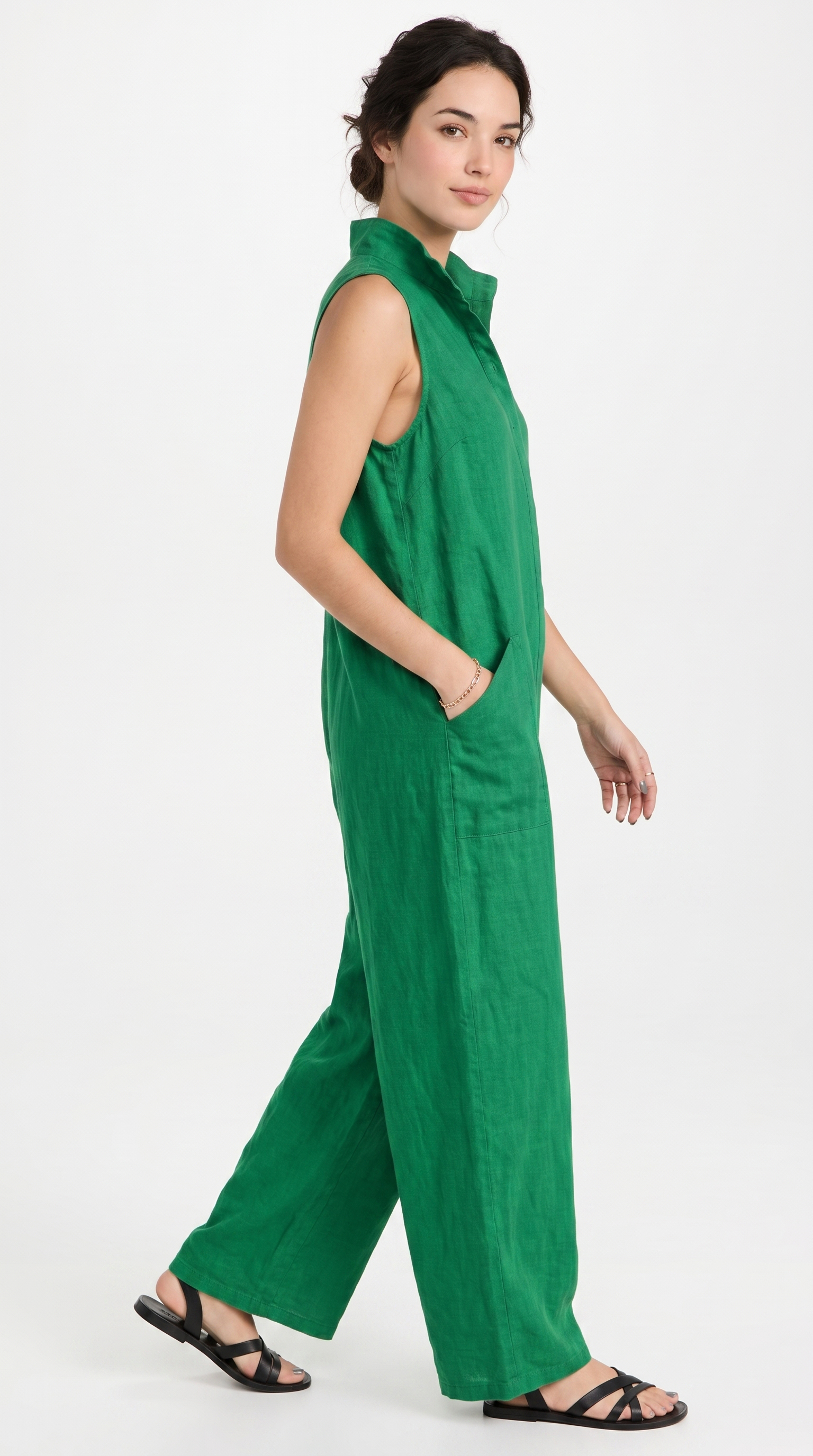A woman modeling a sleeveless green wide-leg jumpsuit with a high collar and side pockets, paired with black strappy sandals, against a plain white studio background.