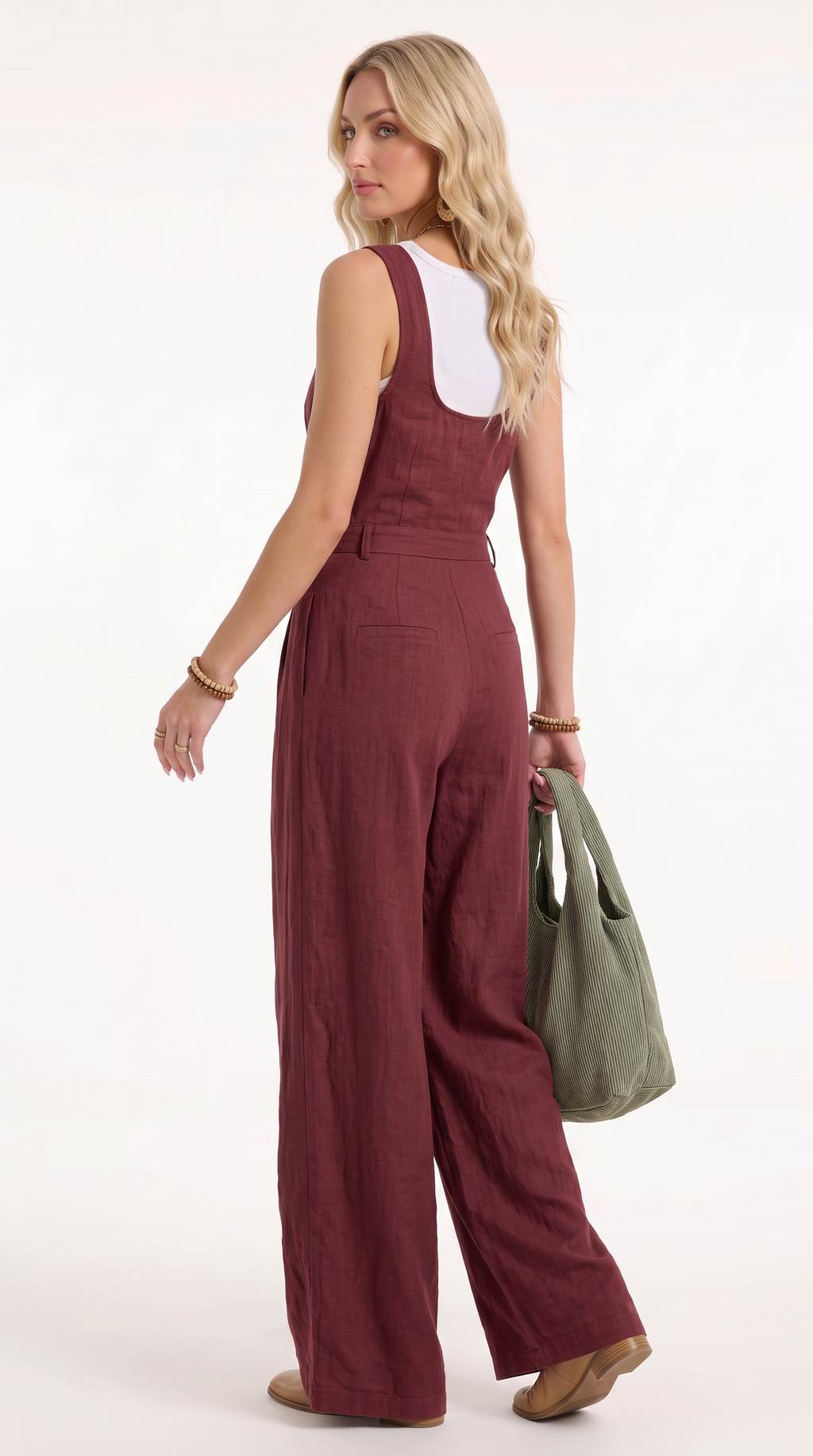 A woman modeling a sleeveless wide-leg jumpsuit in burgundy, shown from a back-three-quarter angle. She is holding an olive green tote bag and wearing a white undershirt, gold bracelets, and tan shoes against a plain white studio background.
