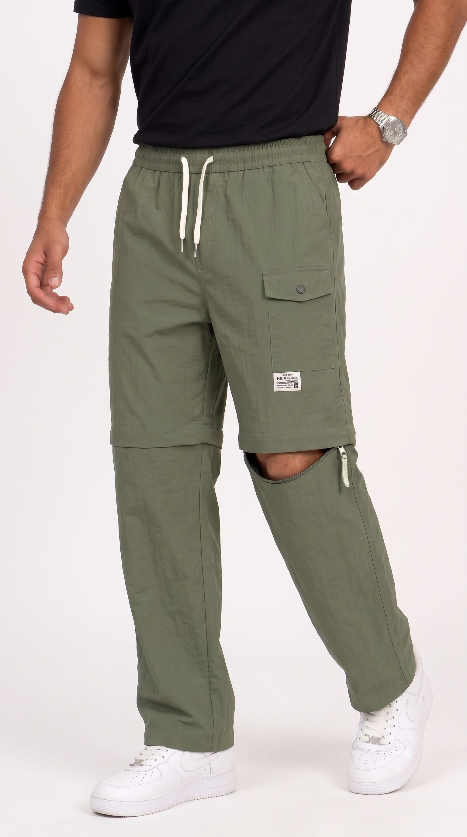 A person wearing olive green cargo-style pants with a drawstring waist, flap cargo pocket, and zippered leg openings, paired with a black shirt and white sneakers against a plain light background, from Meleche Fashion USA or Meléche (short as (Mé))