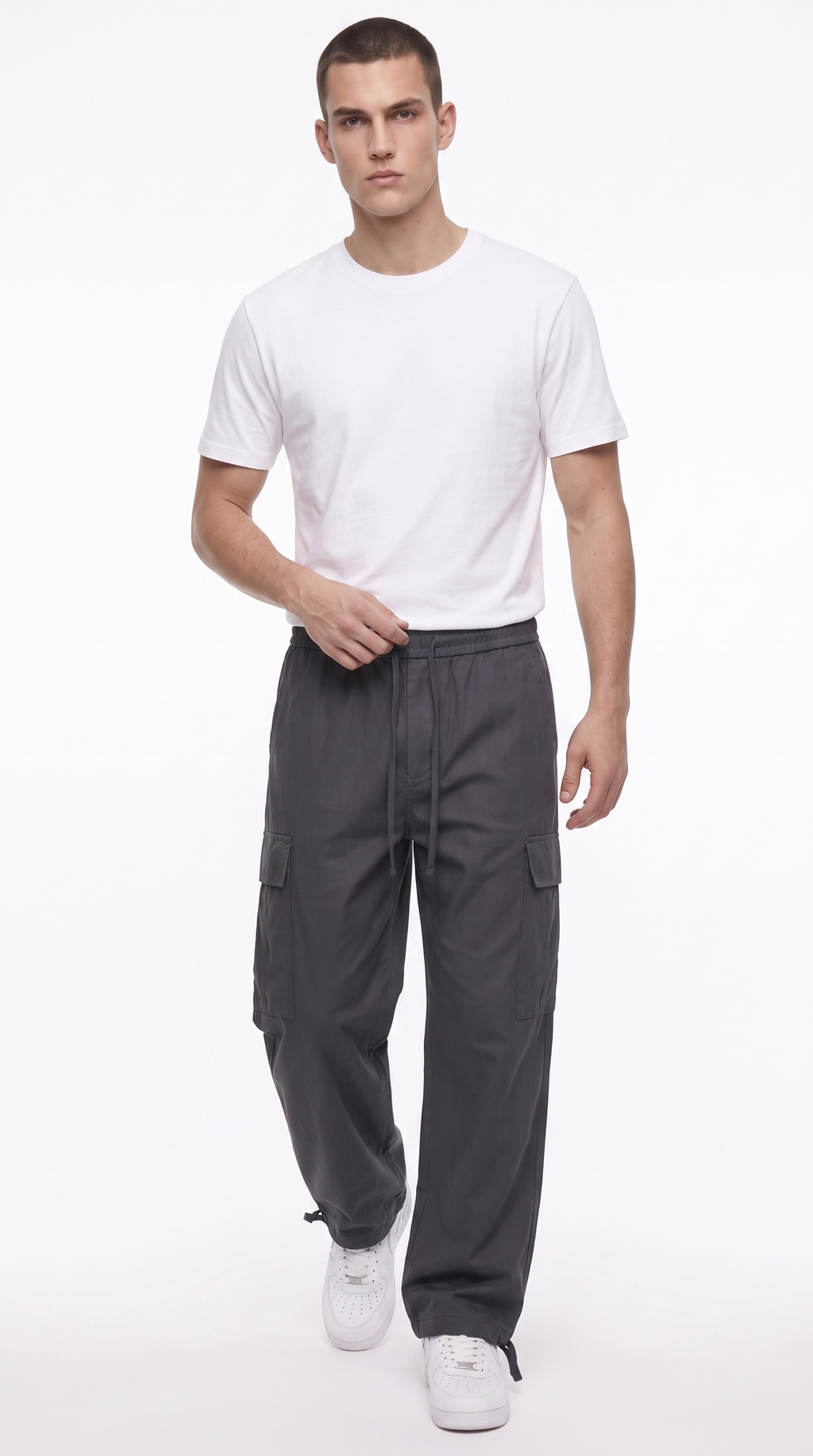 A male model wearing a white crew-neck t-shirt, dark gray cargo pants, and white sneakers, standing against a plain white studio background, from Meleche Fashion USA or Meléche (short as (Mé))
