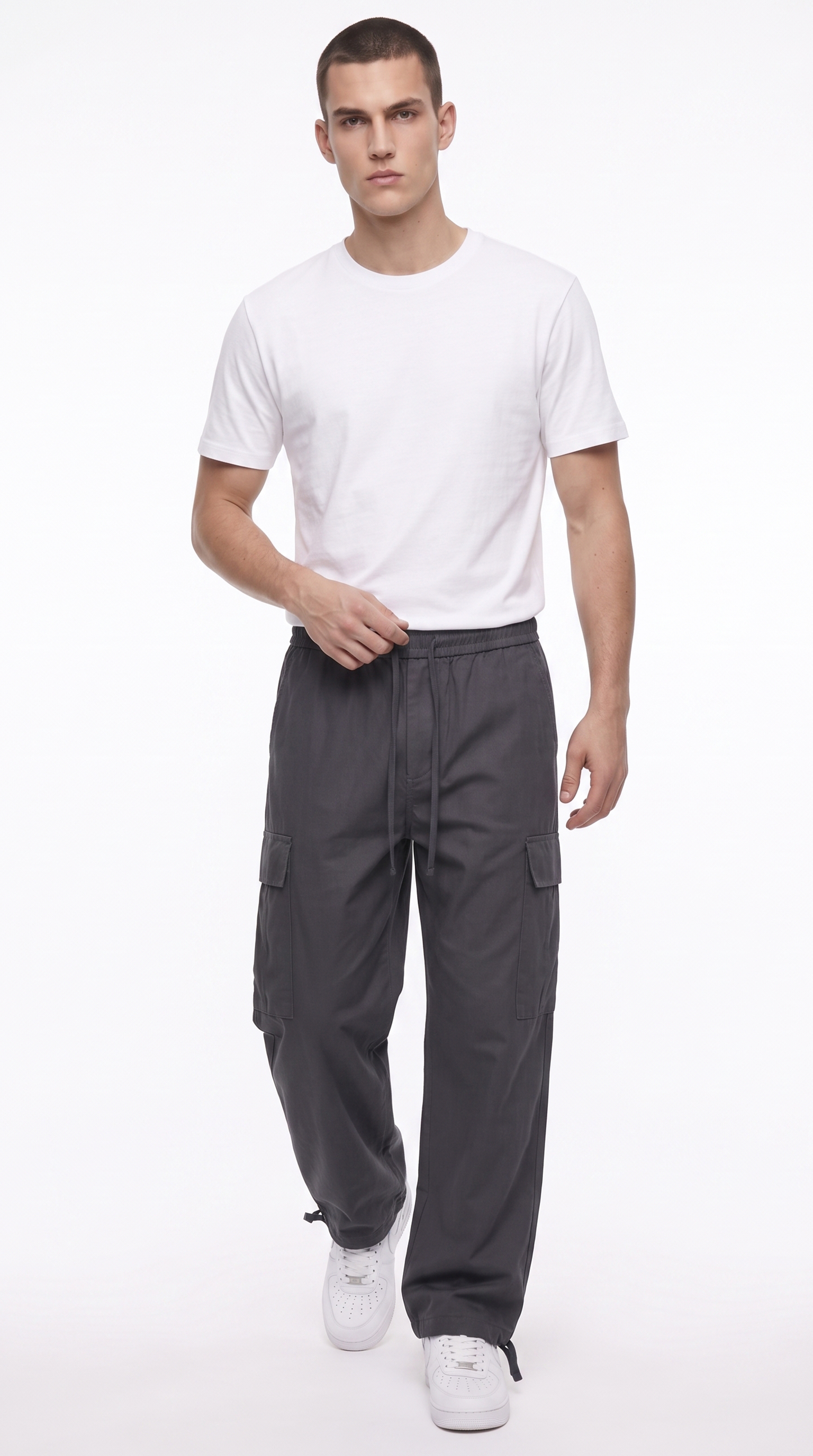 A male model wearing a white crew-neck t-shirt, dark gray cargo pants, and white sneakers, standing against a plain white studio background, from Meleche Fashion USA or Meléche (short as (Mé))