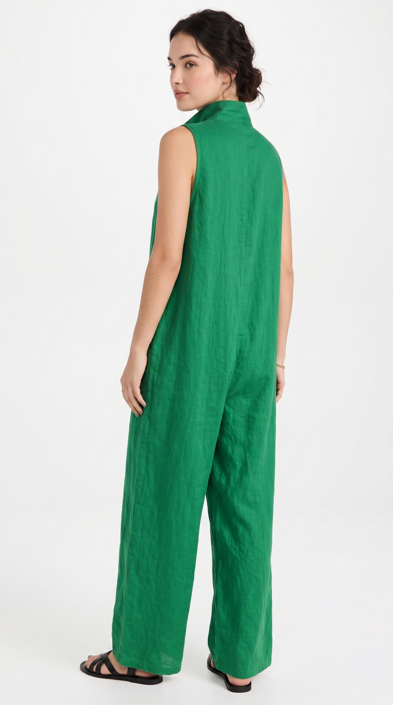 A woman wearing a bright green sleeveless jumpsuit with a high mock neck and wide legs, shown from a three-quarters back view against a plain white studio background.