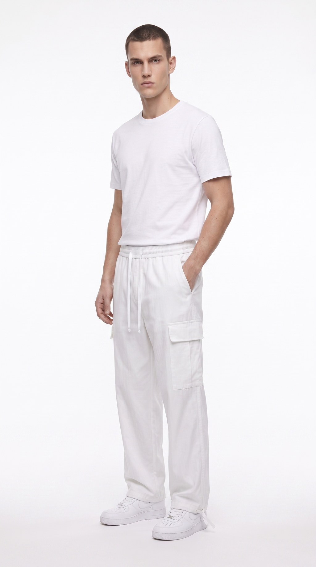 A man modeling a minimalist white outfit consisting of a short-sleeve crewneck t-shirt, loose-fit cargo pants with a drawstring waist, and white sneakers, set against a plain white studio background, from Meleche Fashion USA or Meléche (short as (Mé))