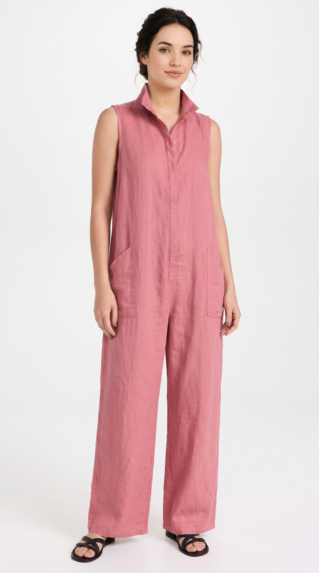 A woman modeling a sleeveless dusty pink linen jumpsuit with a button-front placket and wide legs, paired with black sandals, against a plain white studio background.