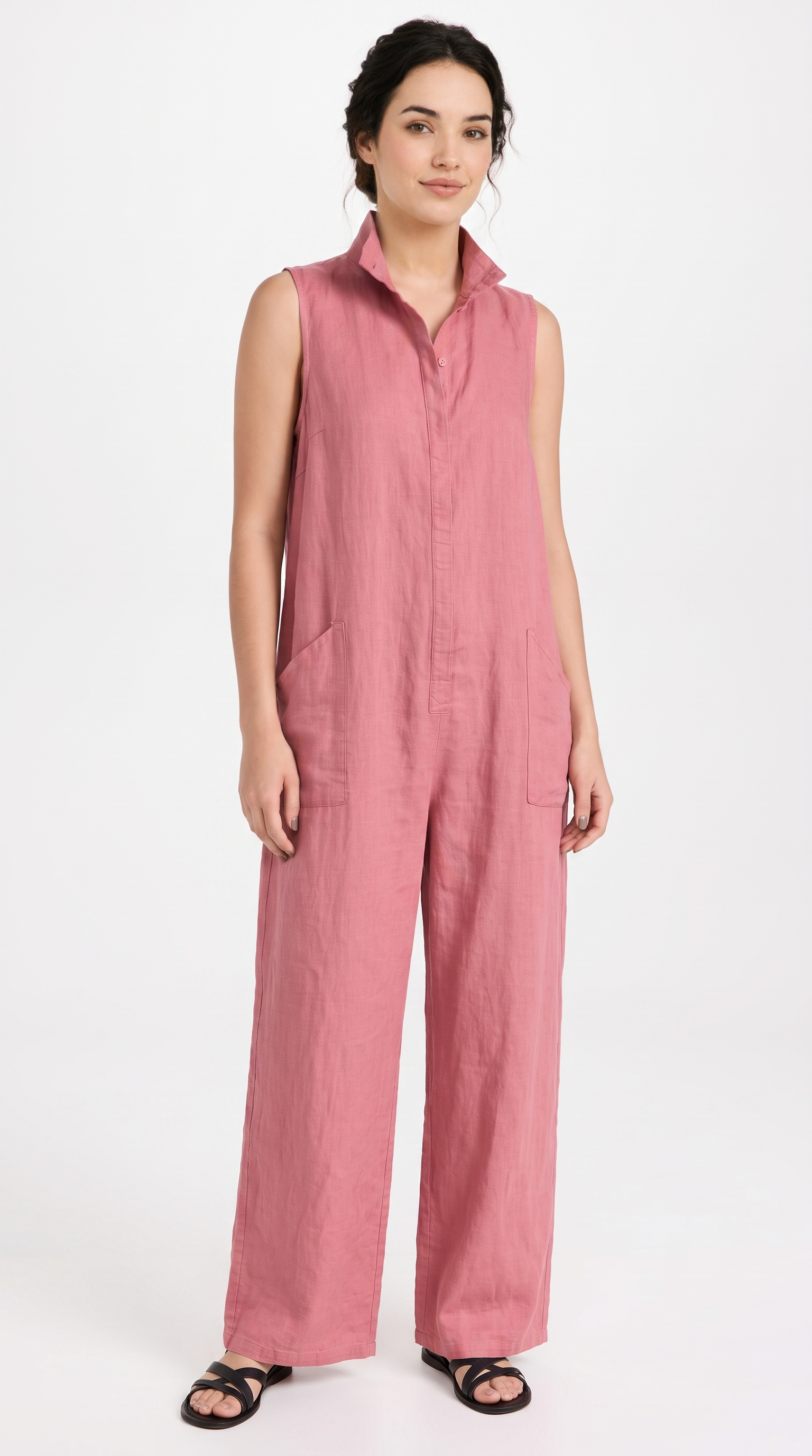 A woman modeling a sleeveless dusty pink linen jumpsuit with a button-front placket and wide legs, paired with black sandals, against a plain white studio background.