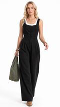 A woman modeling a black sleeveless wide-leg jumpsuit with a white scoop neck trim, holding an olive green tote bag against a plain white background.