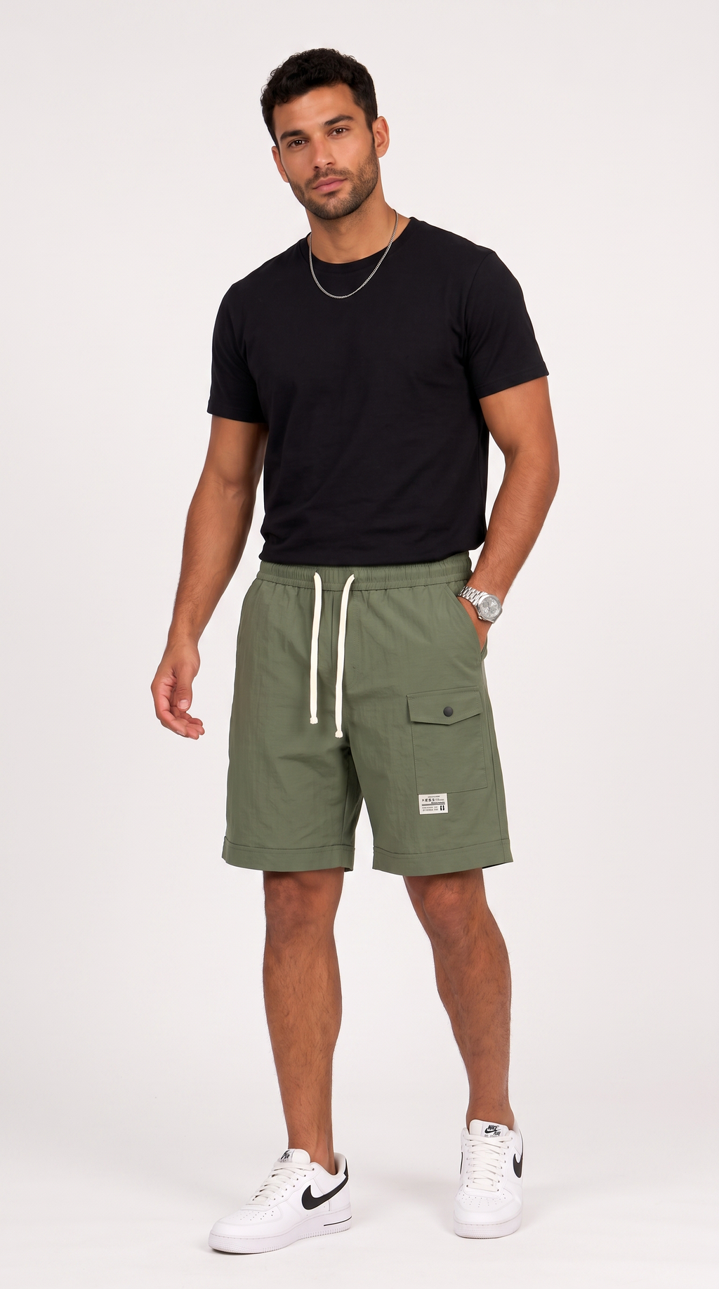 A man modeling olive green cargo shorts with white drawstrings, paired with a black t-shirt and white sneakers, standing against a light gray studio background, from Meleche Fashion USA or Meléche (short as (Mé))