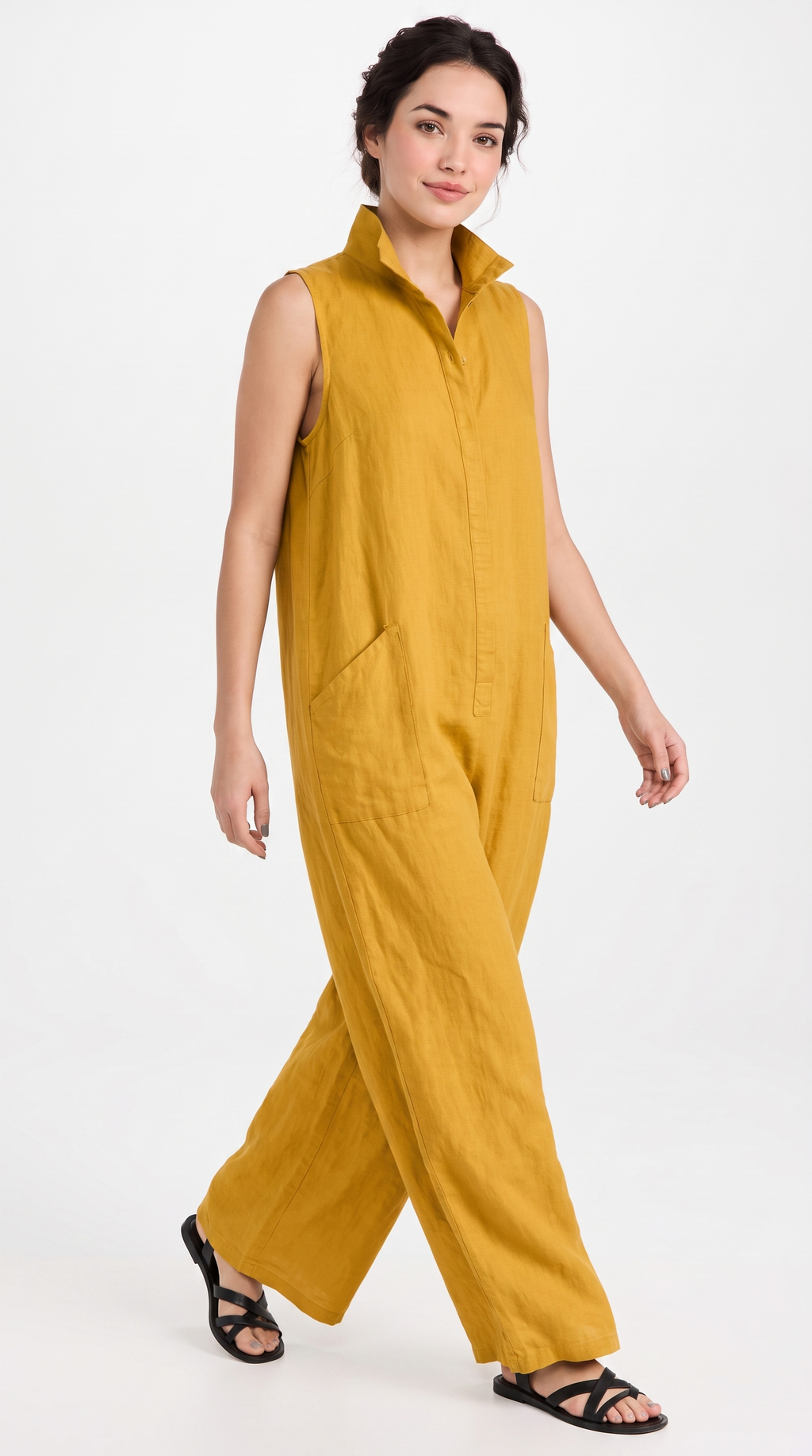 A woman modeling a sleeveless mustard yellow linen jumpsuit with a collar and wide legs, paired with black strappy sandals, against a plain white studio background.