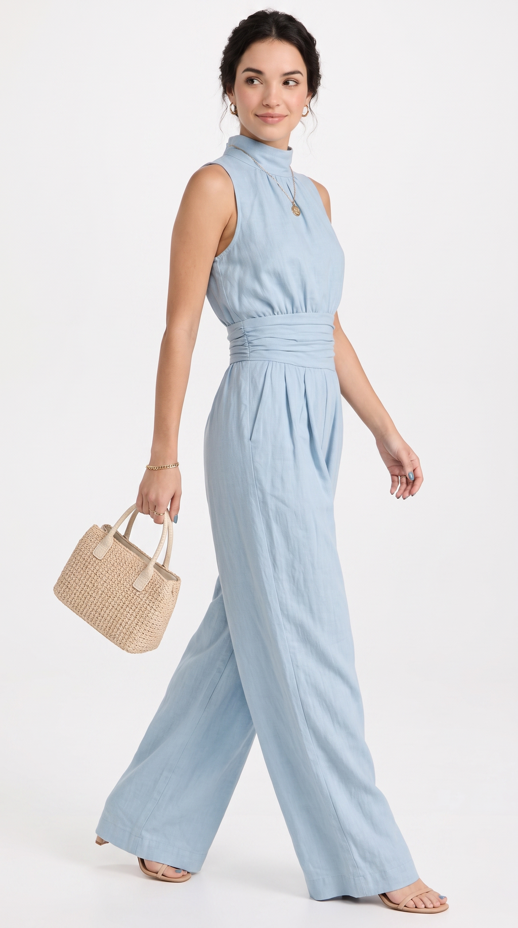 A woman modeling a light blue sleeveless jumpsuit with a high mock neck, gathered waist, and wide legs, holding a small woven straw handbag against a plain white studio background, from Meleche Fashion USA or Meléche (short as (Mé))
