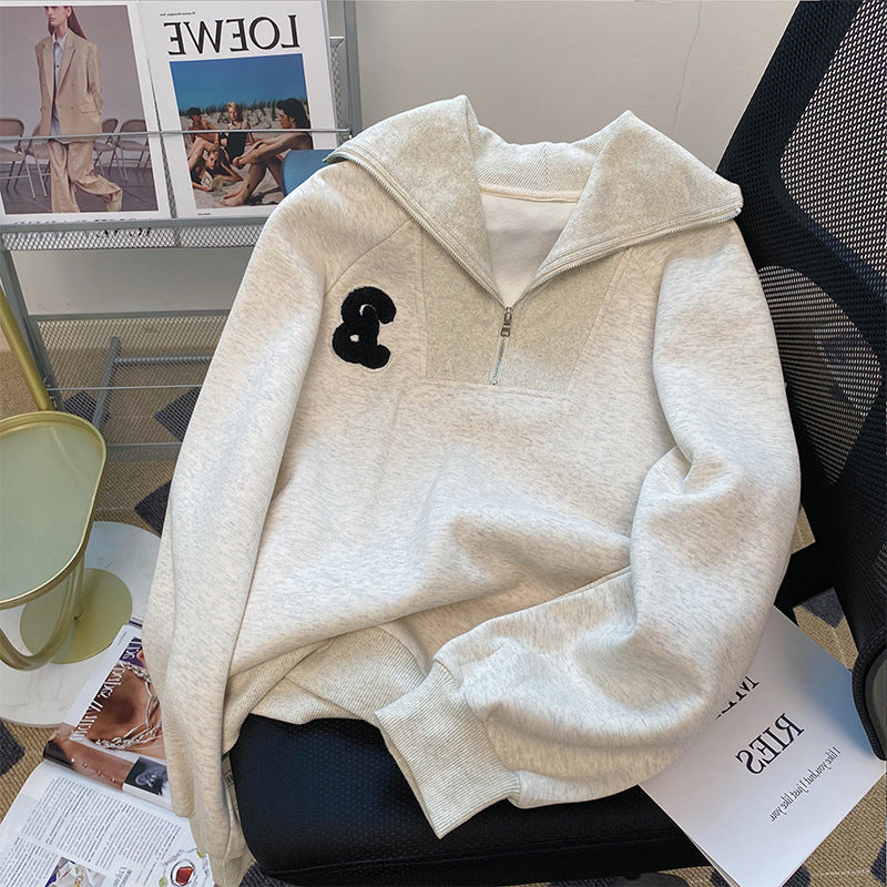 Beige half-zip hoodie with a black logo on a chair with magazines underneath