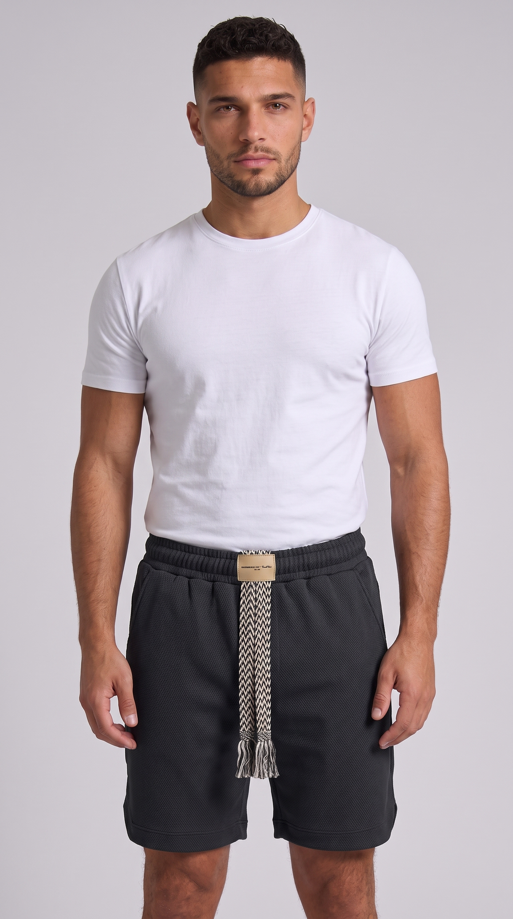 A man modeling a white short-sleeve crewneck t-shirt and dark charcoal gray shorts with a patterned tie belt, shot against a solid light gray background, from Meleche Fashion USA or Meléche (short as (Mé))