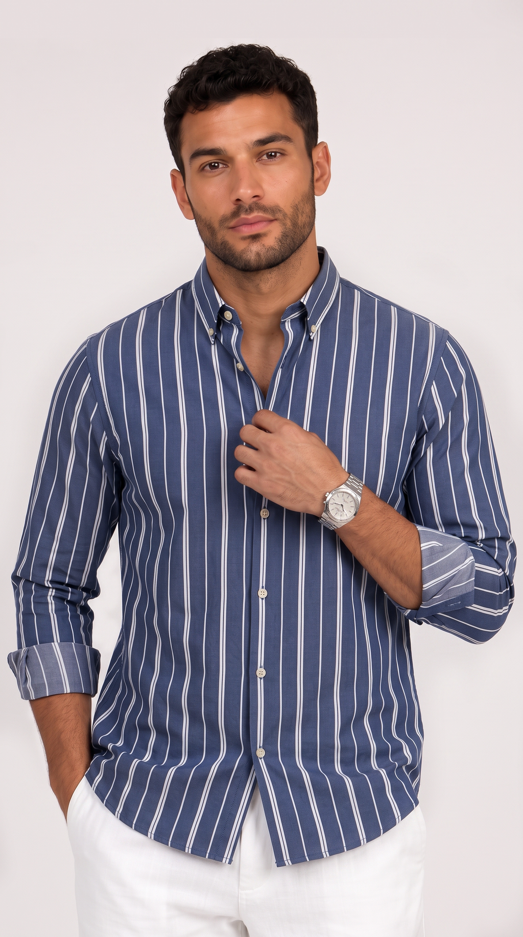 A man wearing a blue and white vertically striped long-sleeve button-down shirt with white trousers, standing against a light gray studio background, from Meleche Fashion USA or Meléche (short as (Mé))