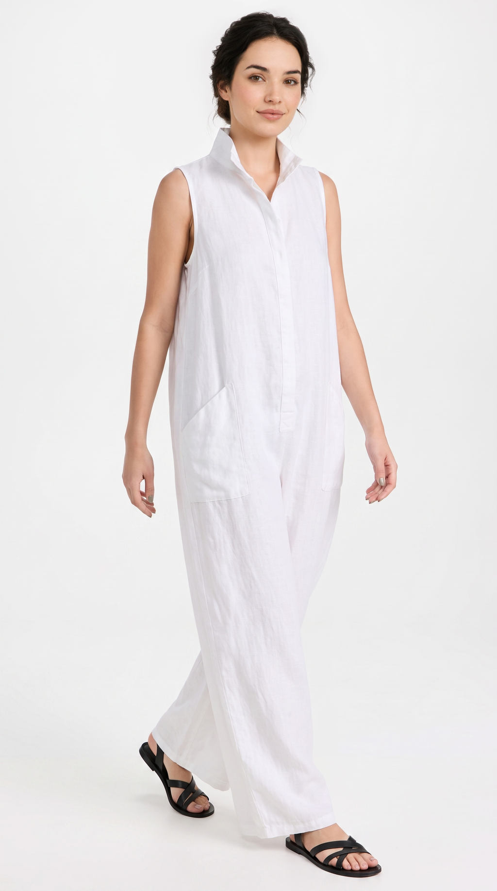 A woman modeling a white sleeveless collared jumpsuit with a button-front bodice and wide legs, paired with black strappy sandals, against a plain white studio background.