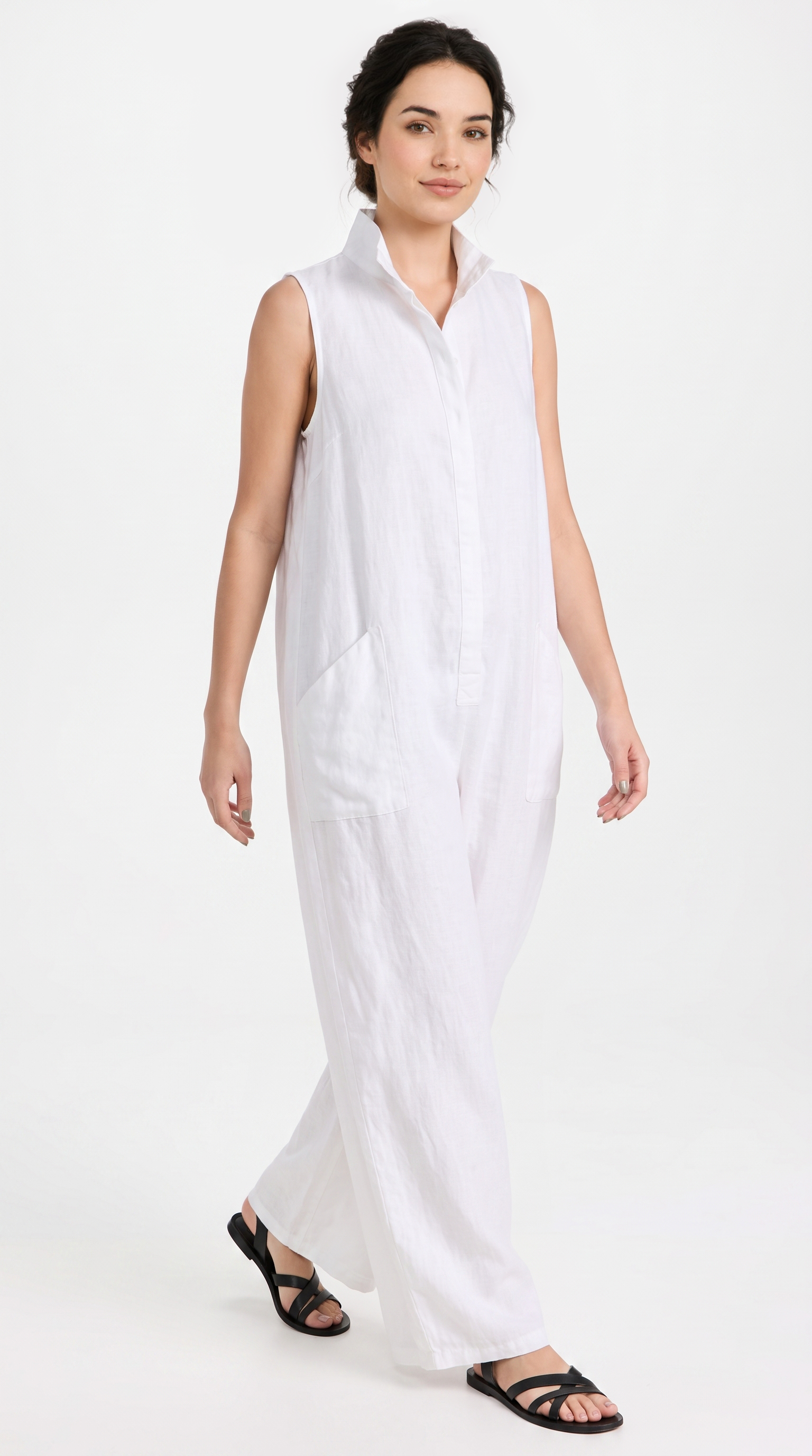A woman modeling a white sleeveless collared jumpsuit with a button-front bodice and wide legs, paired with black strappy sandals, against a plain white studio background.