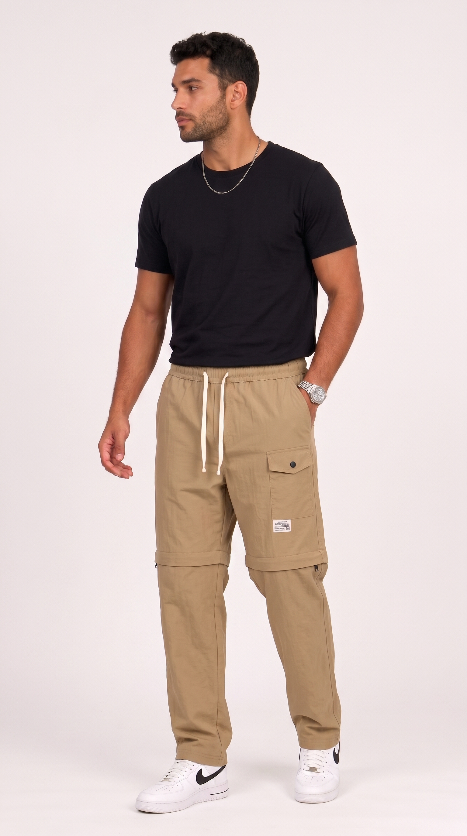 A man modeling a pair of beige cargo jogger pants with white drawstrings, paired with a black t-shirt and white sneakers, standing against a light gray studio background, from Meleche Fashion USA or Meléche (short as (Mé))