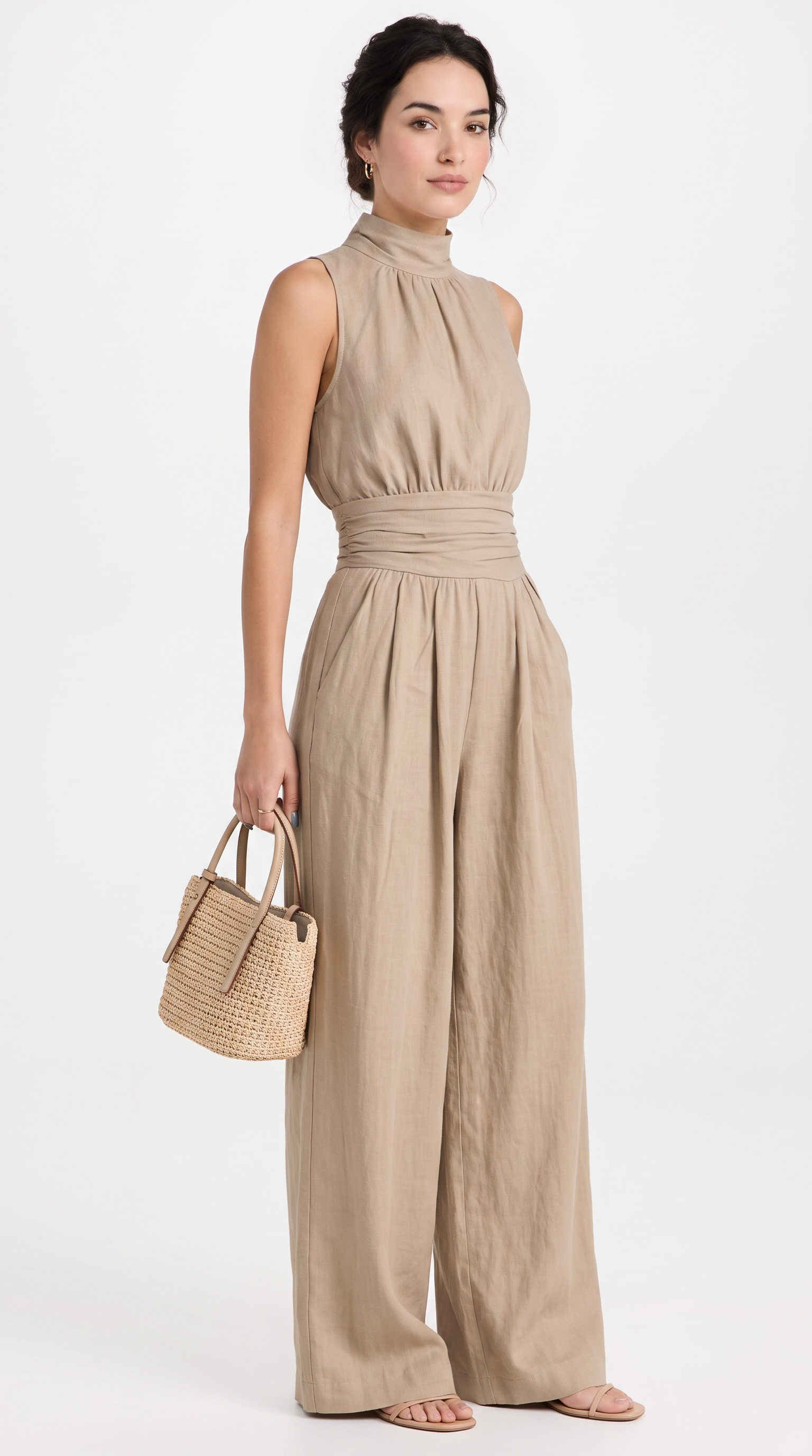 A woman modeling a sleeveless, high-neck, wide-leg jumpsuit in a light beige linen-like fabric, holding a small woven straw handbag against a plain white studio background, from Meleche Fashion USA or Meléche (short as (Mé))