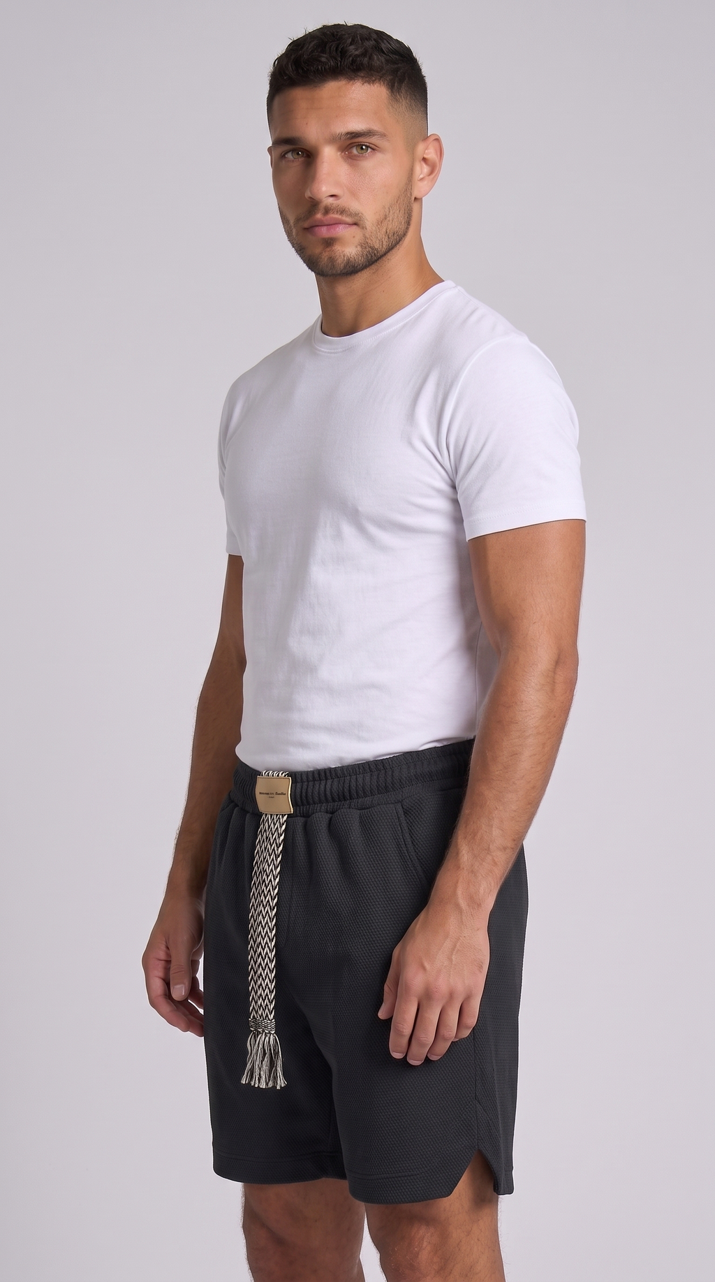 A man modeling a white short-sleeve crewneck t-shirt and dark gray athletic shorts with a decorative braided belt, standing against a light gray studio background, from Meleche Fashion USA or Meléche (short as (Mé))