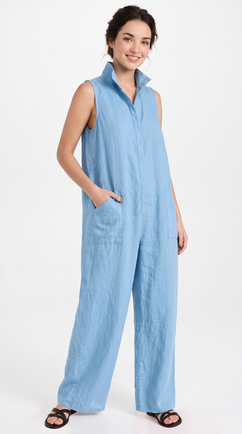 A woman modeling a light blue sleeveless wide-leg jumpsuit with a collared neckline and front pockets, paired with black sandals, against a plain white studio background.