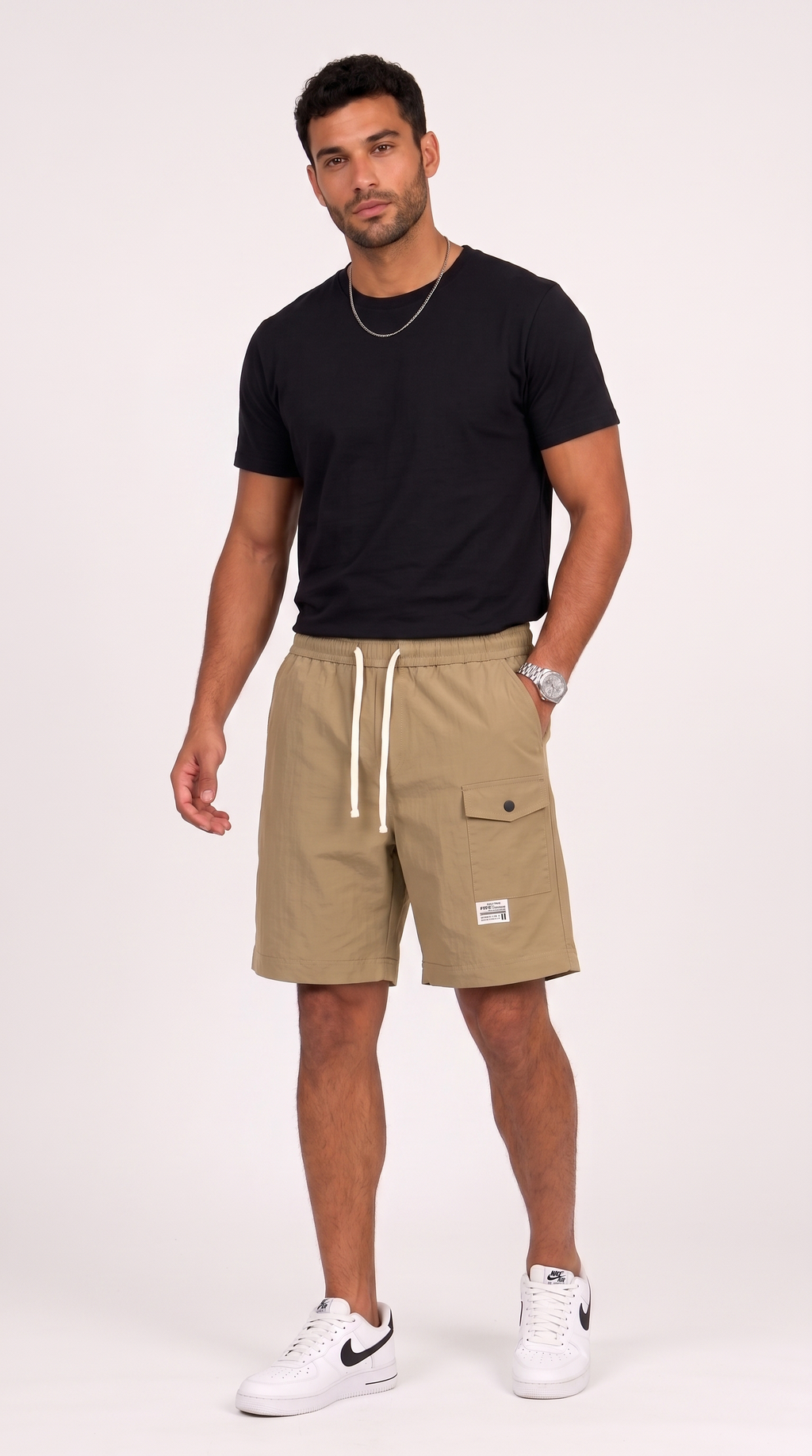 A man modeling khaki cargo shorts with a white drawstring, paired with a black t-shirt and white sneakers, standing against a light off-white studio background, from Meleche Fashion USA or Meléche (short as (Mé))