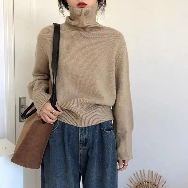 Person wearing a beige turtleneck sweater and blue jeans, holding a brown coat.