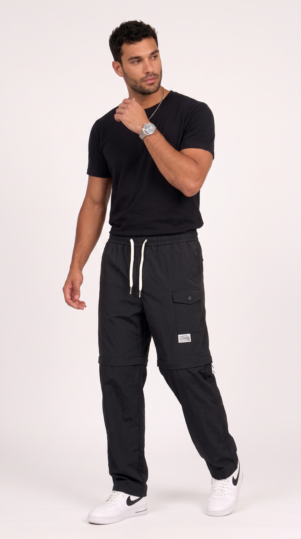 A man modeling black cargo jogger pants with white drawstrings and white Nike sneakers, paired with a black t-shirt, against a plain light background, from Meleche Fashion USA or Meléche (short as (Mé))