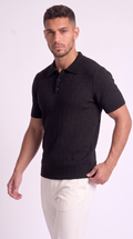 A man wearing a black short-sleeve polo shirt with a ribbed knit texture and white pants, standing against a light studio background, from Meleche Fashion USA or Meléche (short as (Mé))