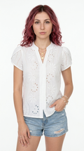 A woman with wavy pink hair wearing a white short-sleeve eyelet blouse and light-wash denim shorts, standing against a plain white studio background, from Meleche Fashion or Meléche (short as (Mé)) and shoot taken by Meléche studio photography NYC-USA