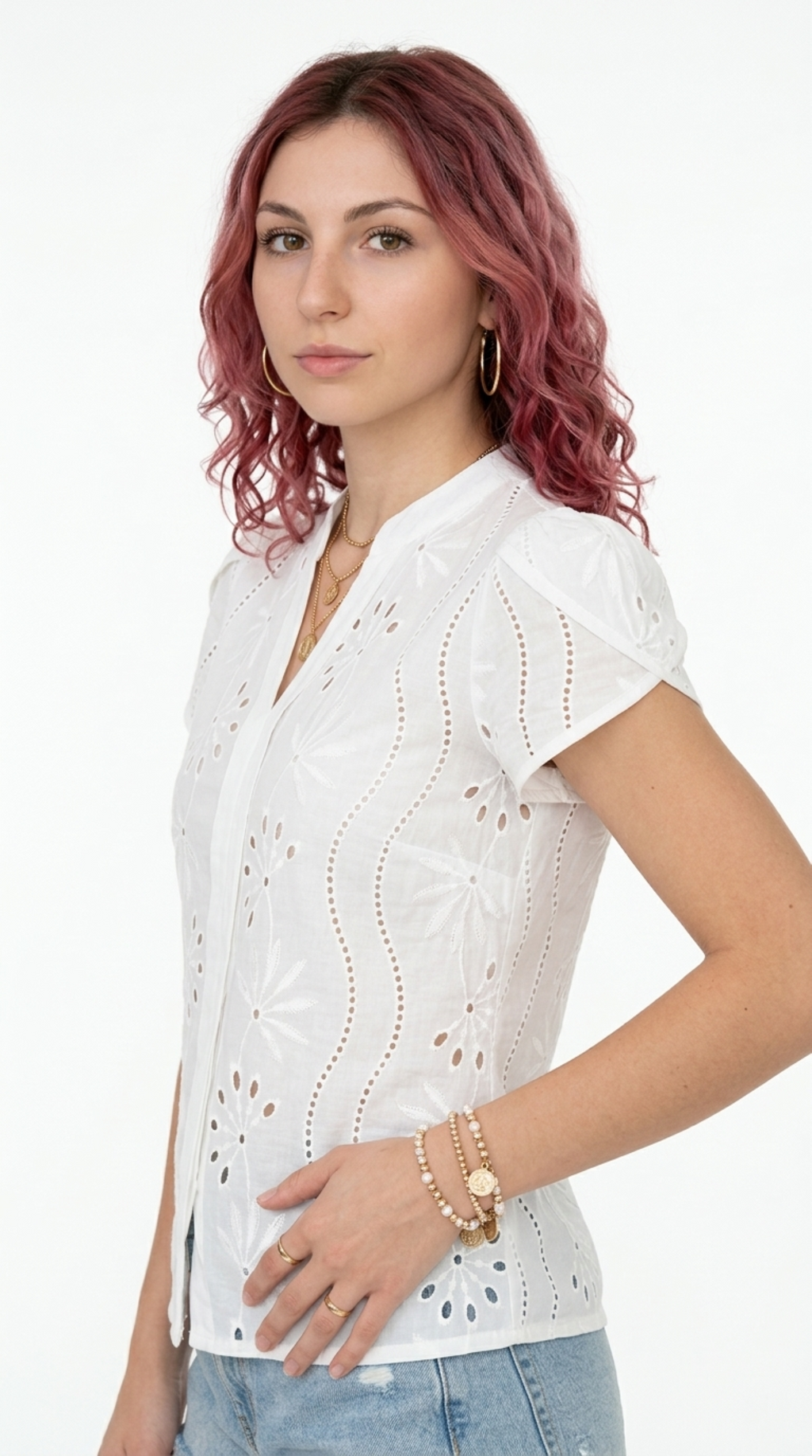 A woman with pinkish-red hair wearing a white short-sleeve eyelet blouse and light blue jeans, standing against a plain white studio background, from Meleche Fashion or Meléche (short as (Mé)) and shoot taken by Meléche studio photography NYC-USA