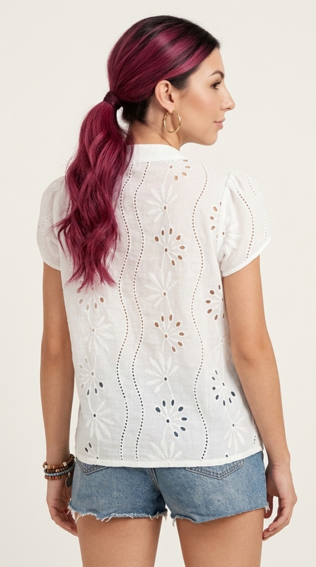 A woman shown from the back wearing a white short-sleeve eyelet blouse with vertical floral cutout patterns and light blue denim shorts, against a plain light background, from Meleche Fashion or Meléche (short as (Mé)) and shoot taken by Meléche studio photography NYC-USA