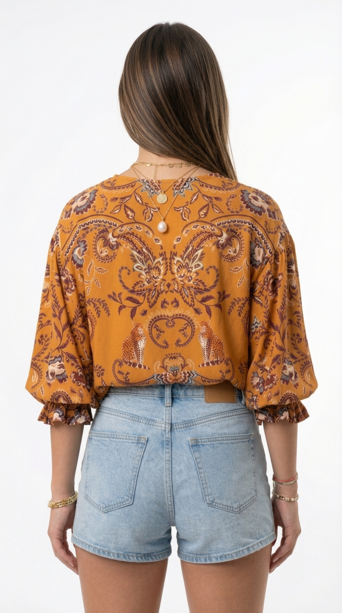 A woman shown from the back wearing a mustard yellow patterned blouse with ruffled sleeves and light blue denim shorts against a white studio background, from Meleche Fashion or Meléche (short as (Mé)) and shoot taken by Meléche studio photography NYC-USA