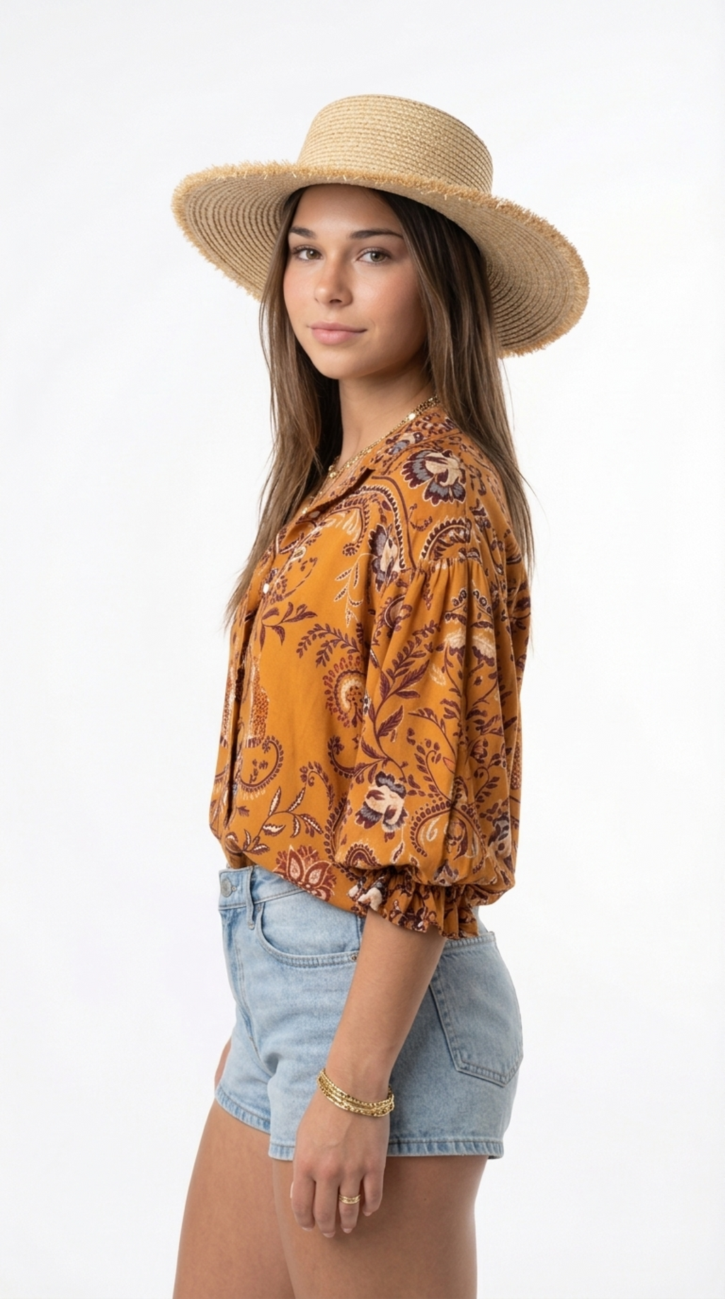 A woman wearing a straw sun hat, a mustard yellow patterned blouse with brown paisley details, and light blue denim shorts, photographed against a plain white studio background, from Meleche Fashion or Meléche (short as (Mé)) and shoot taken by Meléche studio photography NYC-USA