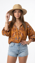 A woman modeling a mustard yellow bohemian patterned blouse with ruffled sleeves, paired with denim shorts and a straw hat, against a plain white background, from Meleche Fashion or Meléche (short as (Mé)) and shoot taken by Meléche studio photography NYC-USA