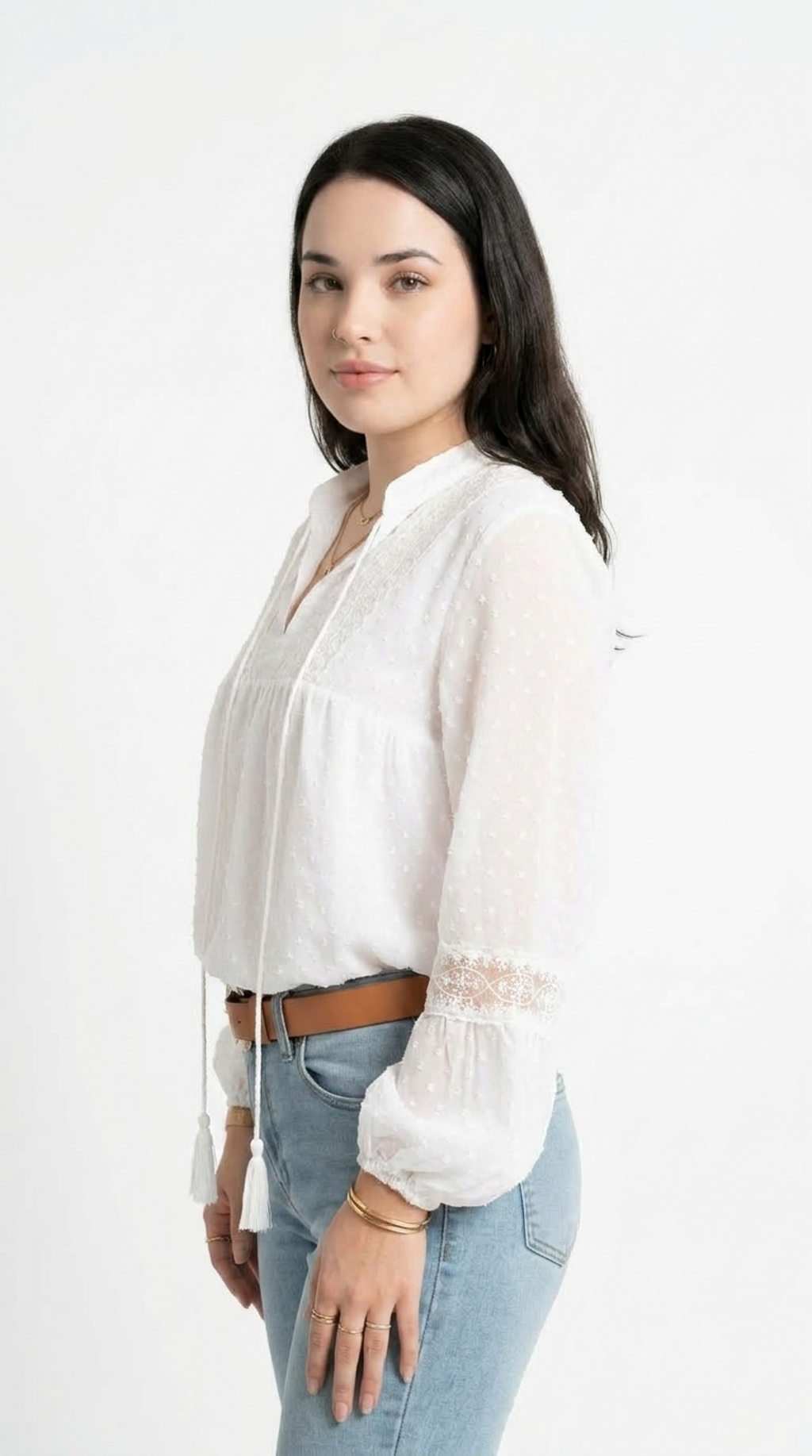 A woman wearing a white long-sleeve blouse with lace trim and tassel ties, paired with light blue jeans and a brown belt, standing against a plain white background, from Meleche Fashion USA or Meléche (short as (Mé))