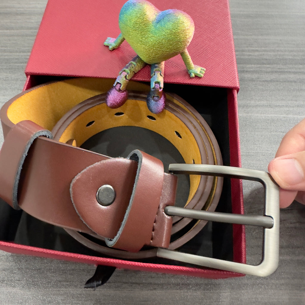 Gael belt Brown leather belt with silver buckle on a gray surface, colorful frog toy on red box from Meleche or Meléche (short as (Mé))