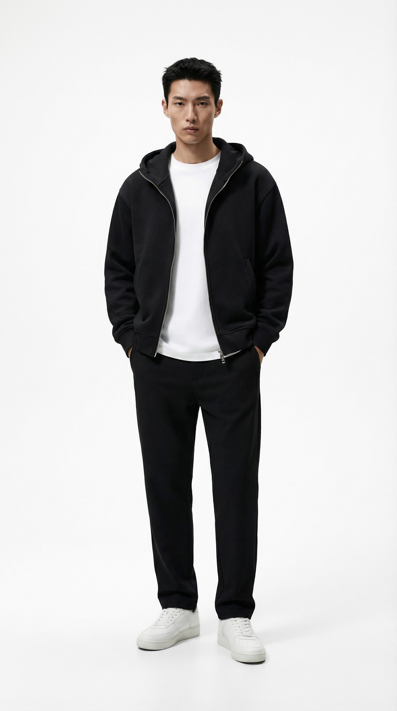 Person wearing a black jacket, white shirt, black pants, and white sneakers against a plain wall.