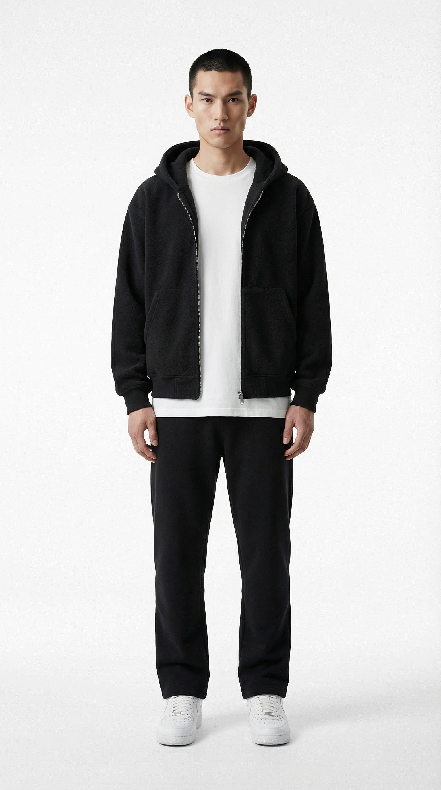 Person wearing a black jacket, white shirt, black pants, and white shoes against a gray concrete wall.