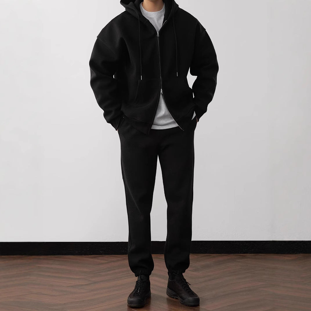 Person wearing a black hoodie, pants, and boots standing against a plain white wall.